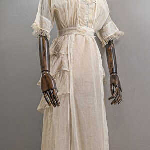 May include: A white sheer dress with a ruffled hem and a sash at the waist. The dress is displayed on a wooden mannequin with brown arms.