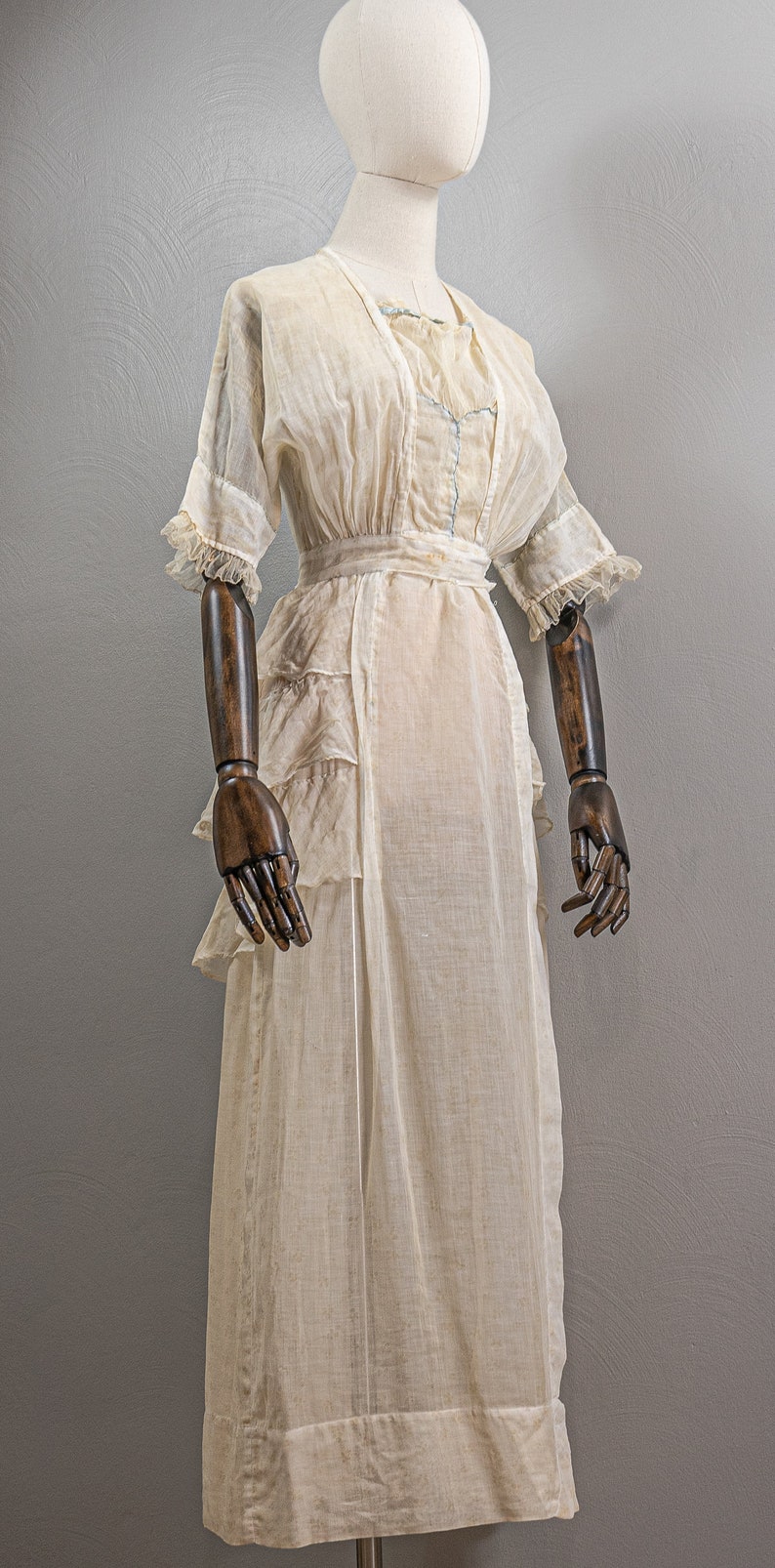 May include: A white sheer dress with a ruffled hem and a sash at the waist. The dress is displayed on a wooden mannequin with brown arms.