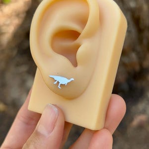 May include: A light blue dinosaur stud earring is displayed on a tan ear model. The earring is a small, detailed silhouette of a dinosaur. The background is blurred, suggesting an outdoor setting.