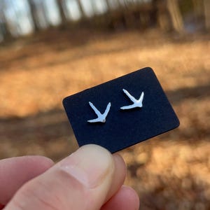 May include: Silver-colored bird footprint stud earrings displayed on a black card. The earrings are held up against a blurred background of trees and foliage. The earrings are small and delicate, perfect for everyday wear.