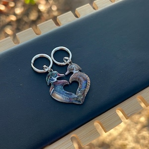 May include: A metal charm featuring two wood ducks facing each other, their bodies curved to form a heart shape. The ducks display detailed plumage in shades of brown, green, white, and blue. Two silver-toned metal rings are attached to the top.