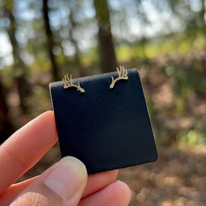 May include: A pair of gold-toned stud earrings shaped like deer antlers. The earrings are mounted on a small, square, black textured display card. The metal has a smooth, polished finish, and each earring features the distinct branching points of antlers.