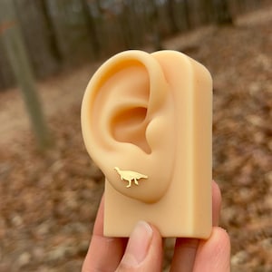 May include: A pair of gold-colored dinosaur stud earrings displayed on a flesh-toned ear model. The earrings are small and feature a silhouette of a dinosaur. The model is held in a hand against a blurred outdoor background.