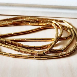 Könnte beinhalten: Nahaufnahme von mehreren Strängen goldfarbener, zylindrischer Perlen. Die Perlen sind in einem lockeren, fließenden Muster vor einem hellen Hintergrund angeordnet. Die Perlen scheinen aus Metall zu sein und haben eine glänzende Oberfläche.