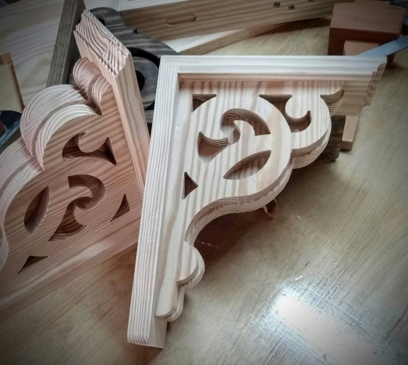 Op de afbeelding: Twee houten hoekbeugels met ingewikkelde uitgesneden ontwerpen. De beugels zijn gemaakt van licht hout en hebben een natuurlijke afwerking.
