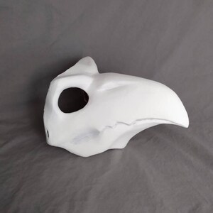 May include: A white, unpainted, plastic mask with a long, curved beak. The mask has a single eye hole and a small hole on the side.