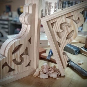 Op de afbeelding: Twee onafgewerkte houten hoekbeugels met ingewikkelde uitgesneden ontwerpen. De beugels zijn gemaakt van licht hout en hebben een natuurlijke afwerking. Houtsnippers zijn verspreid rond de beugels.