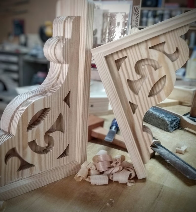 Op de afbeelding: Twee onafgewerkte houten hoekbeugels met ingewikkelde uitgesneden ontwerpen. De beugels zijn gemaakt van licht hout en hebben een natuurlijke afwerking. Houtsnippers zijn verspreid rond de beugels.