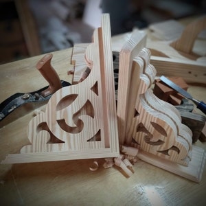 Op de afbeelding: Twee onafgewerkte houten plankdragers met ingewikkelde uitsnijdingen. De dragers zijn gemaakt van licht hout en hebben een natuurlijke afwerking. Ze staan op een houten werkbank met houtkrullen eromheen.