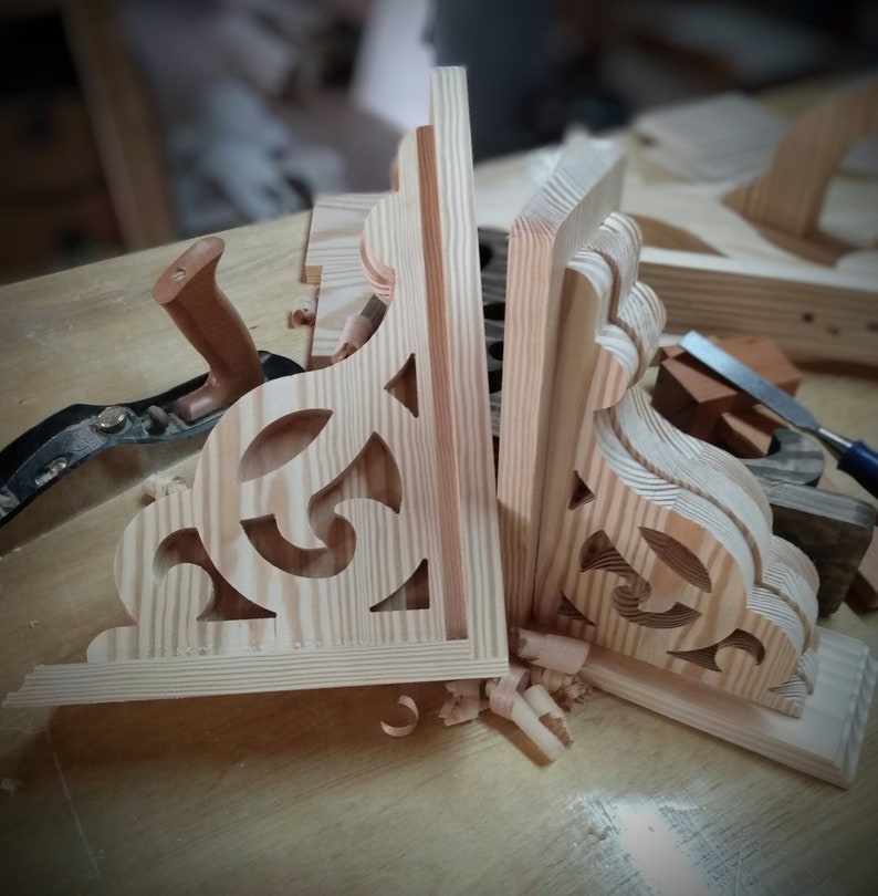 Op de afbeelding: Twee onafgewerkte houten plankdragers met ingewikkelde uitsnijdingen. De dragers zijn gemaakt van licht hout en hebben een natuurlijke afwerking. Ze staan op een houten werkbank met houtkrullen eromheen.