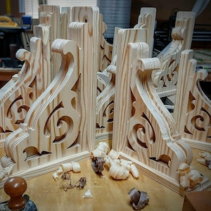 May include: Several unfinished wooden corbels with ornate scrollwork and cut-out patterns. The corbels are made of light-coloured wood and are arranged on a wooden surface, with wood shavings scattered nearby.