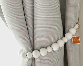 May include: White wooden bead curtain tieback with a brown leather strap. The tieback is holding back a pair of gray striped curtains. The tieback has a small brown tag with the text "Decoazul" on it.