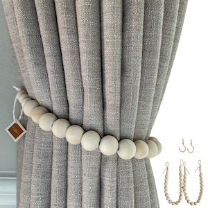 May include: A light gray curtain is held back by a wooden bead tieback. Additional wooden bead tiebacks and metal hooks are displayed to the right. The curtain has a textured appearance. The wooden beads are light in color.