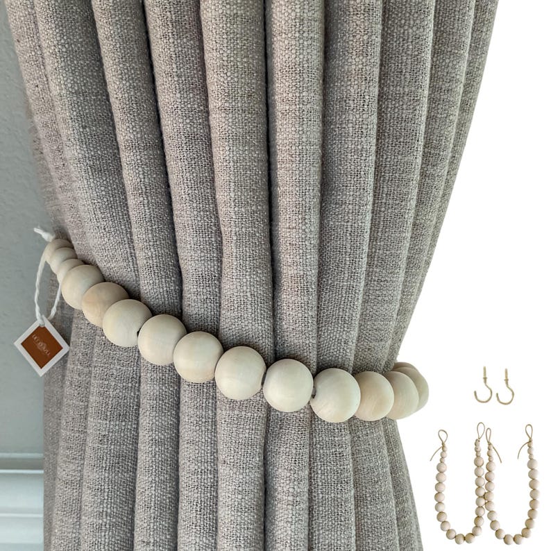 May include: A light gray curtain is held back by a wooden bead tieback. Additional wooden bead tiebacks and metal hooks are displayed to the right. The curtain has a textured appearance. The wooden beads are light in color.