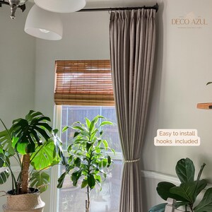 May include: Taupe-colored curtain panel with a beaded tieback, hanging in front of a window with a bamboo shade. The text "Easy to install hooks included" is visible. The room features potted plants and a ceiling light.