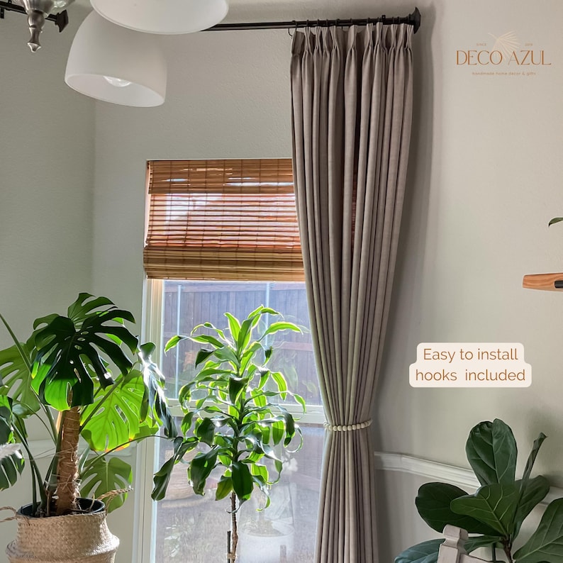 May include: Taupe-colored curtain panel with a beaded tieback, hanging in front of a window with a bamboo shade. The text "Easy to install hooks included" is visible. The room features potted plants and a ceiling light.