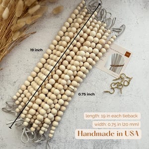 May include: Wooden bead curtain tiebacks, each 19 inches long and 0.75 inches wide. The tiebacks are made of light-colored wooden beads and have gray fabric loops. The image also shows a small card and gold-colored metal hooks. Handmade in the USA.