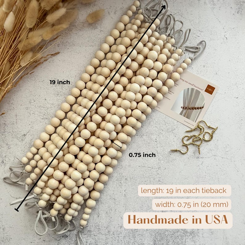 May include: Wooden bead curtain tiebacks, each 19 inches long and 0.75 inches wide. The tiebacks are made of light-colored wooden beads and have gray fabric loops. The image also shows a small card and gold-colored metal hooks. Handmade in the USA.