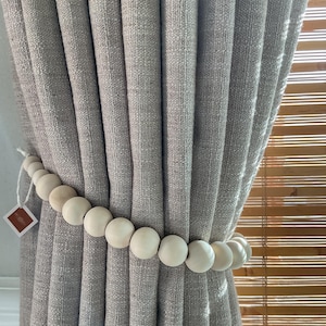 May include: A light gray curtain is held back by a beaded curtain tieback. The tieback is made of round, light-colored wooden beads. A small tag is attached to the tieback.
