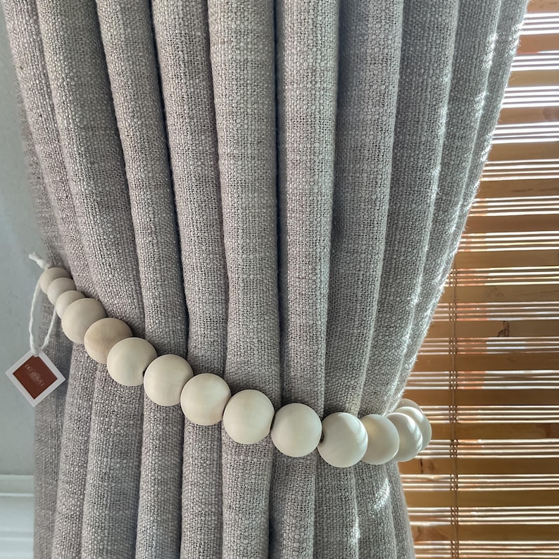 May include: A light gray curtain is held back by a beaded curtain tieback. The tieback is made of round, light-colored wooden beads. A small tag is attached to the tieback.