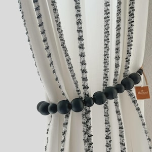 May include: White and black striped curtain panels held by a black beaded curtain tieback. The tieback is made of various sized round beads. A small brown tag with the text "DecoAzul" is attached to the tieback.