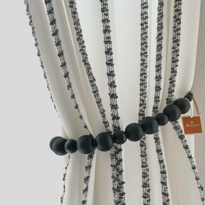 May include: White and black striped curtain panels held by a black beaded curtain tieback. The tieback is made of various sized round beads. A small brown tag with the text "DecoAzul" is attached to the tieback.