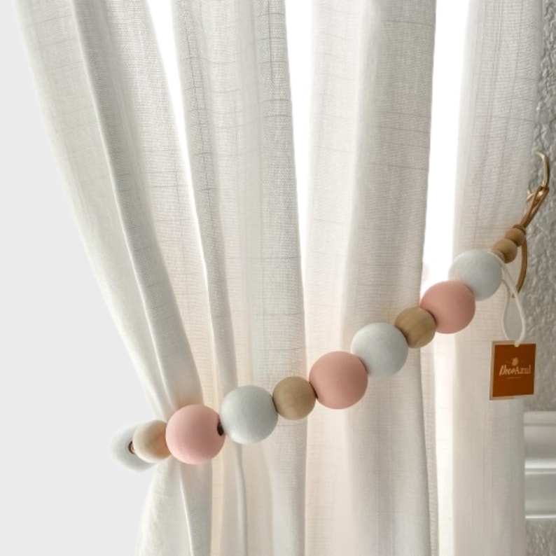 May include: A beaded curtain tieback with alternating white, pink, and natural wood beads. The tieback is holding back a white curtain. A small tag is attached to the tieback.