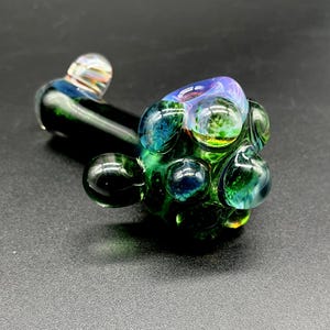 May include: A hand pipe crafted from dark green glass, featuring a bulbous bowl adorned with multiple raised, translucent glass spheres in shades of blue, green, and iridescent purple. The stem is smooth and dark green with a clear glass accent.