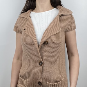 Könnte beinhalten: Ein braunes Strickjäckchen mit kurzen Ärmeln, einem Kragen und vier Knöpfen auf der Vorderseite. Das Jäckchen hat zwei Taschen auf der Vorderseite.