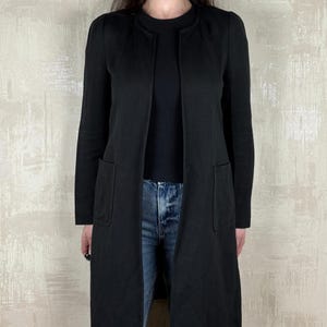 May include: A long black open-front cardigan with two front pockets. The cardigan is worn over a black top and blue jeans. The cardigan has long sleeves and a simple, elegant design, suitable for layering.