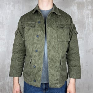 Op de afbeelding: Groene en bruine camouflagejas met vier voorzakken en knoopsluitingen. De jas heeft een kraag en lange mouwen. Het patroon bestaat uit verticale lijnen. De jas wordt gedragen over een grijs t-shirt en een blauwe jeans.