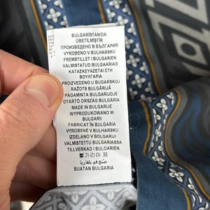May include: Close-up of a patterned fabric with a label. The fabric features a geometric design in shades of blue, grey, and white, with gold accents. The label displays text in multiple languages, including English.