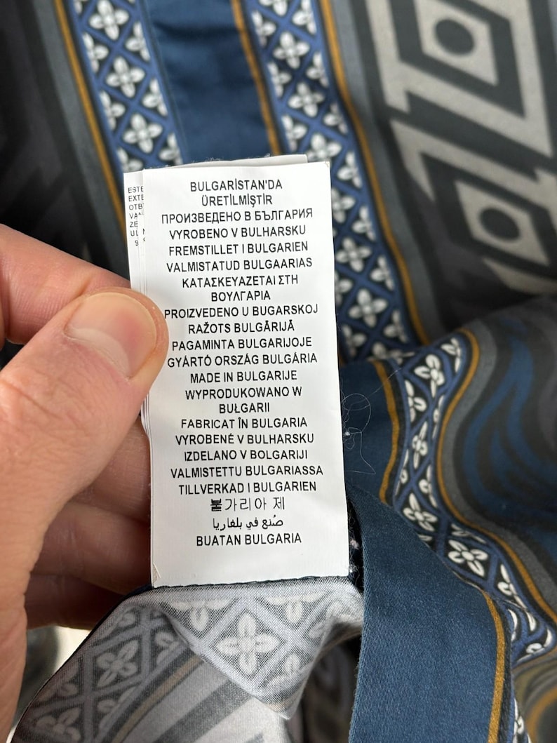 May include: Close-up of a patterned fabric with a label. The fabric features a geometric design in shades of blue, grey, and white, with gold accents. The label displays text in multiple languages, including English.