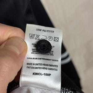 May include: Close-up of a white tag with black text and a black button. The tag reads "100% POLYESTER" and includes washing instructions. The text "MADE IN UNITED ARAB EMIRATES" is also visible. The tag is attached to a black garment.