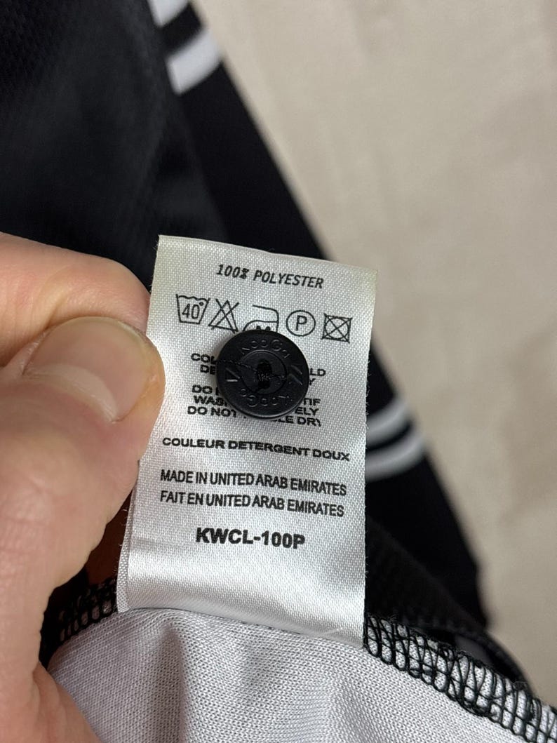 May include: Close-up of a white tag with black text and a black button. The tag reads "100% POLYESTER" and includes washing instructions. The text "MADE IN UNITED ARAB EMIRATES" is also visible. The tag is attached to a black garment.