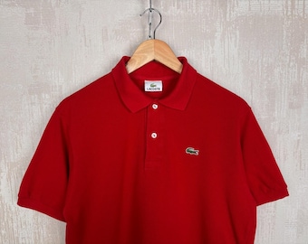 Può includere: Una polo rossa brillante con maniche corte, colletto e abbottonatura a tre bottoni. Il logo Lacoste è visibile sul petto e una piccola etichetta sul colletto. È appesa a una gruccia di legno.