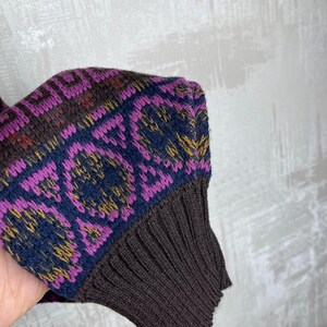 K&ouml;nnte beinhalten: Nahaufnahme eines &Auml;rmels eines Pullovers. Das B&uuml;ndchen ist einfarbig dunkelbraun, w&auml;hrend der obere Teil ein geometrisches Muster in Lila, Marineblau, Braun und Gelb aufweist. Das Strickwerk hat eine gerippte Textur.
