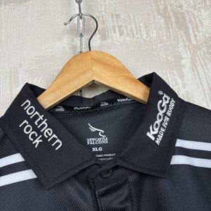May include: Black polo shirt with white stripes and collar details. The shirt features the text "northern rock" and "Newcastle Falcons". The collar also has the text "KooGa MADE FOR RUGBY". The shirt is hanging on a wooden hanger.