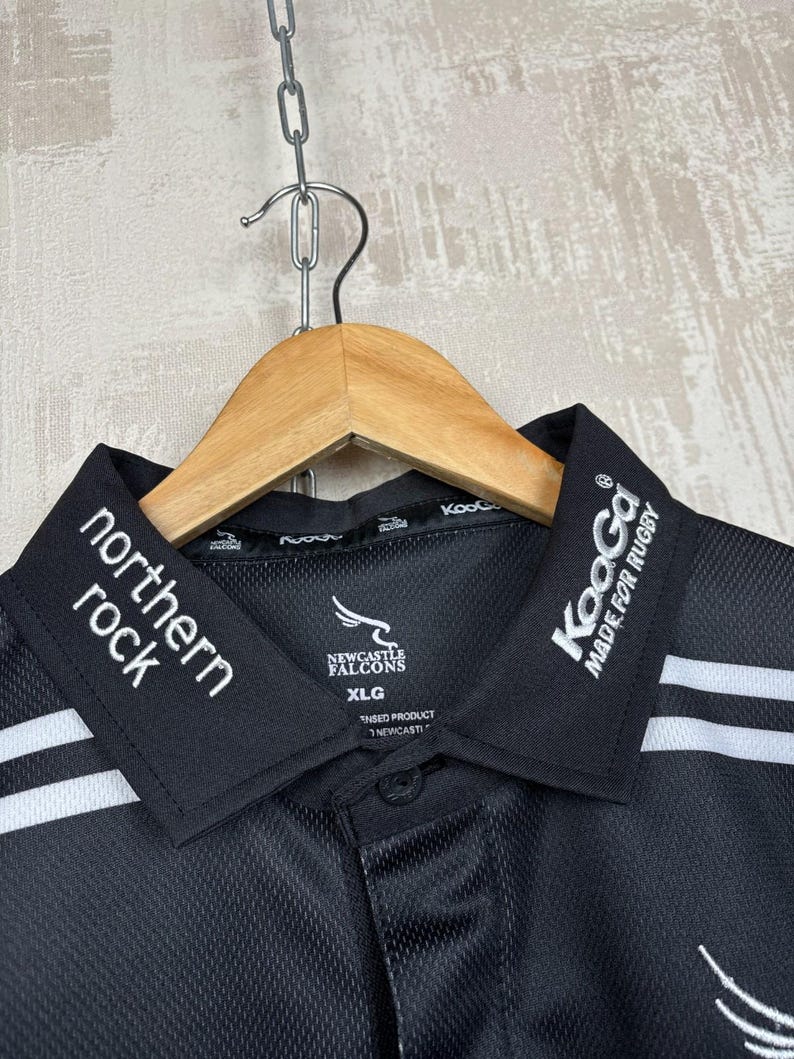 May include: Black polo shirt with white stripes and collar details. The shirt features the text "northern rock" and "Newcastle Falcons". The collar also has the text "KooGa MADE FOR RUGBY". The shirt is hanging on a wooden hanger.
