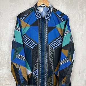 May include: A patterned button-down shirt in blue, teal, brown, and black, hanging on a wooden hanger. The geometric design features diamond and square shapes. The shirt is against a neutral background. The shirt has long sleeves.