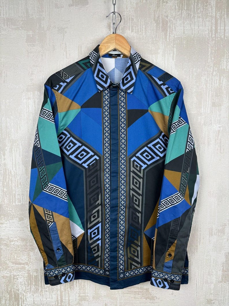 May include: A patterned button-down shirt in blue, teal, brown, and black, hanging on a wooden hanger. The geometric design features diamond and square shapes. The shirt is against a neutral background. The shirt has long sleeves.