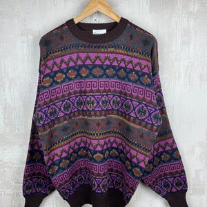 K&ouml;nnte beinhalten: Ein Vintage-Pullover mit geometrischem Muster in Braun-, Lila- und Blaut&ouml;nen. Der Pullover hat einen Rundhalsausschnitt und lange &Auml;rmel. Das Design umfasst Zickzacklinien, Rauten und andere Formen. Der Pullover h&auml;ngt an einem Holzb&uuml;gel.