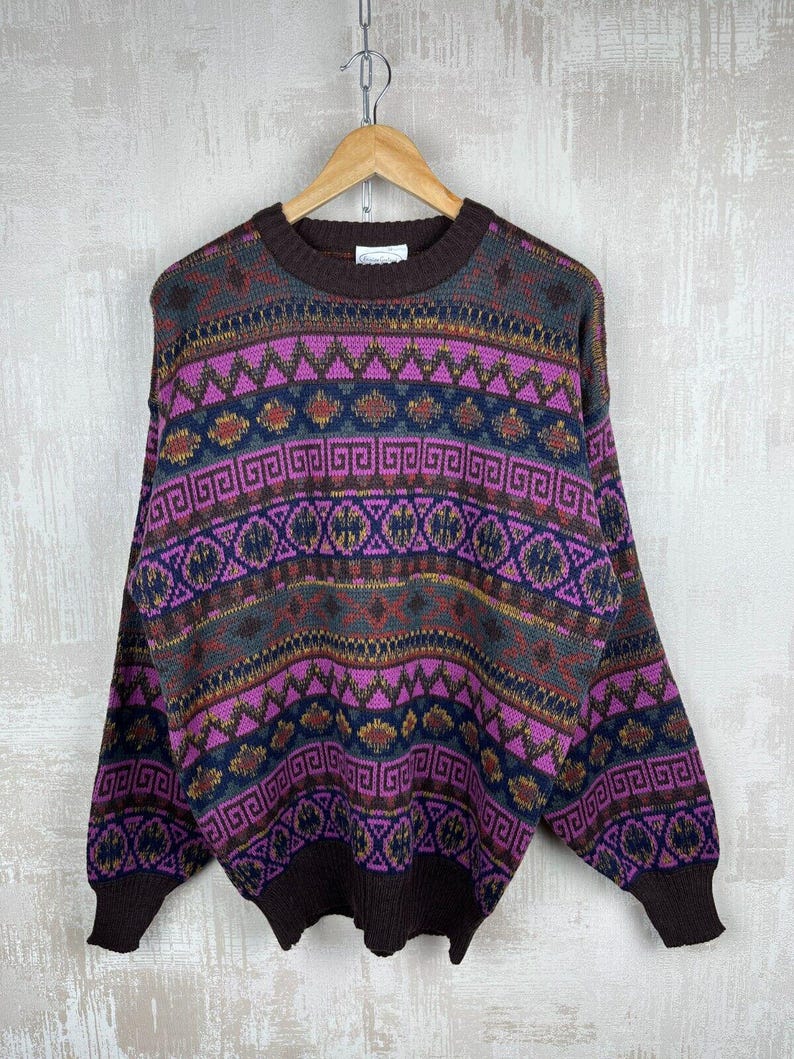 K&ouml;nnte beinhalten: Ein Vintage-Pullover mit geometrischem Muster in Braun-, Lila- und Blaut&ouml;nen. Der Pullover hat einen Rundhalsausschnitt und lange &Auml;rmel. Das Design umfasst Zickzacklinien, Rauten und andere Formen. Der Pullover h&auml;ngt an einem Holzb&uuml;gel.