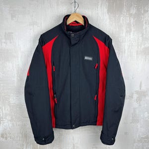 May include: Black and red Ferrari jacket with a high collar and zip pockets. The jacket features red accents on the shoulders and sides, with a small Ferrari logo patch on the chest. The jacket is hanging on a wooden hanger.