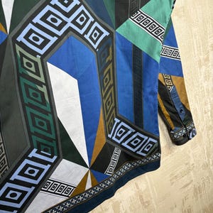 May include: A long-sleeved shirt with a geometric pattern in shades of blue, green, black, white, and gold. The design features angular shapes and repeating square patterns. The shirt has a buttoned cuff and a dark blue border.