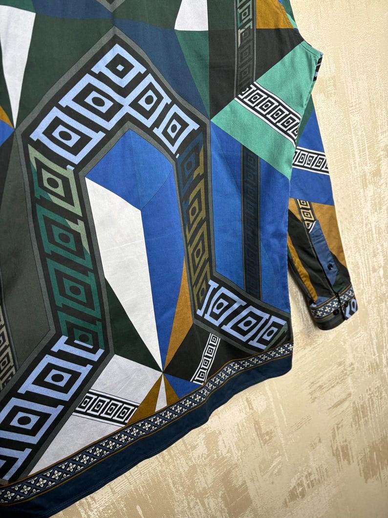 May include: A long-sleeved shirt with a geometric pattern in shades of blue, green, black, white, and gold. The design features angular shapes and repeating square patterns. The shirt has a buttoned cuff and a dark blue border.