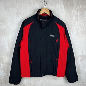 May include: Black and red jacket with a high collar and zippered pockets. The jacket has a black body with red panels on the sides and sleeves. A small rectangular patch with text is on the chest.