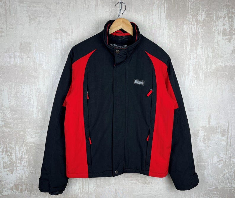 May include: Black and red jacket with a high collar and zippered pockets. The jacket has a black body with red panels on the sides and sleeves. A small rectangular patch with text is on the chest.