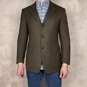Puede incluir: Un blazer verde oliva oscuro con tres botones negros y dos bolsillos delanteros. El blazer se lleva sobre una camisa a cuadros azul y blanca y unos vaqueros azules. La chaqueta tiene un corte clásico y entallado.