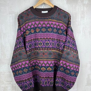 K&ouml;nnte beinhalten: Ein Vintage-Pullover mit Rundhalsausschnitt und geometrischem Muster in Braun-, Lila-, Blau- und Gelbt&ouml;nen. Der Pullover h&auml;ngt an einem Holzb&uuml;gel. Das Design umfasst Zickzack-, Quadrat- und Diamantformen.